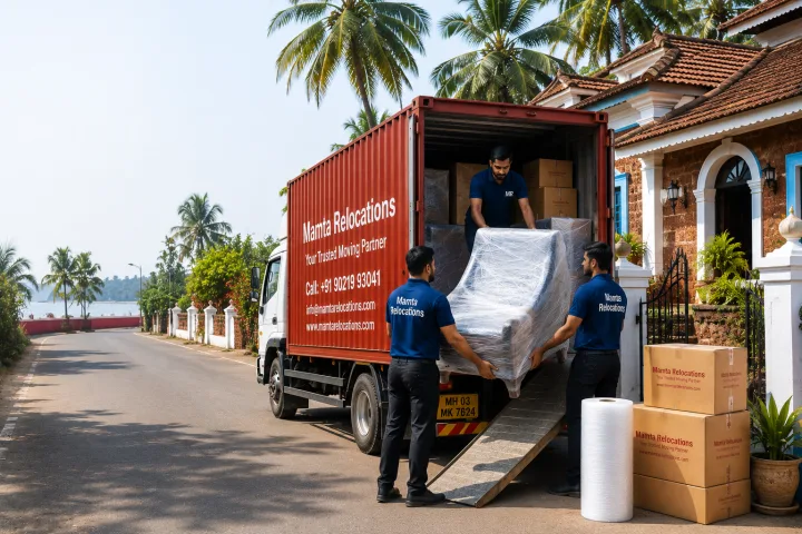 Mamta Relocations crew loading stretch-wrapped furniture and labelled cartons into their truck outside a Portuguese-style laterite house with a