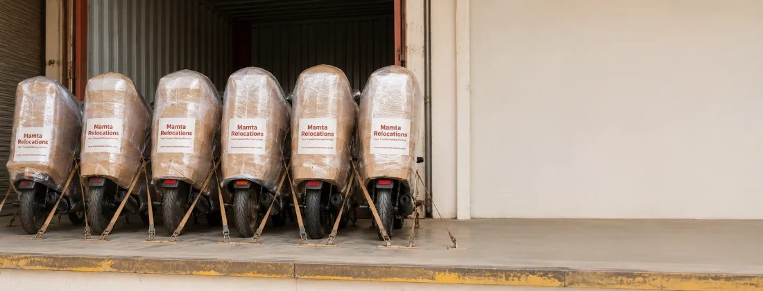 Row of four or five wrapped motorcycles strapped upright and secured side by side inside a loading dock, ready for dispatch — Mamta Relocations