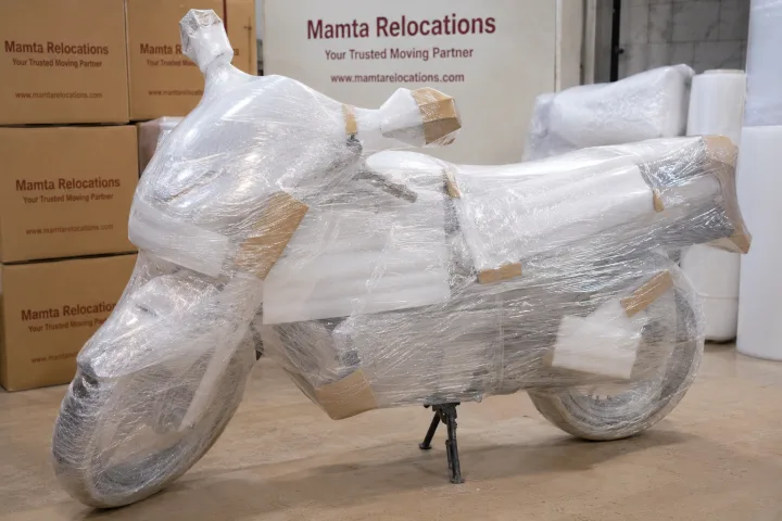 A motorcycle fully cocooned in stretch film and foam padding, mirrors and tank guarded with corner protectors, standing on its centre stand in a