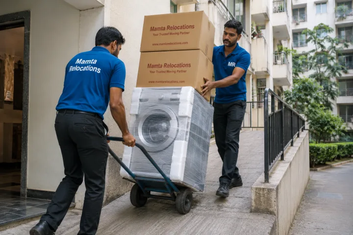 Crew using a sack-barrow trolley to move a stacked washing machine and cartons up an apartment ramp, one worker steadying the load — Mamta