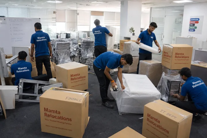 Open-plan office mid-move: crew dismantling and bubble-wrapping workstation desks, office chairs stacked and shrink-filmed, CPUs wrapped
