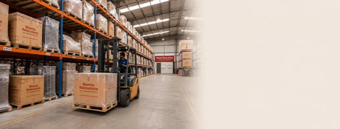 Warehouse interior with long rows of racked, dust-covered household goods and a forklift moving a pallet down the aisle — Mamta Relocations
